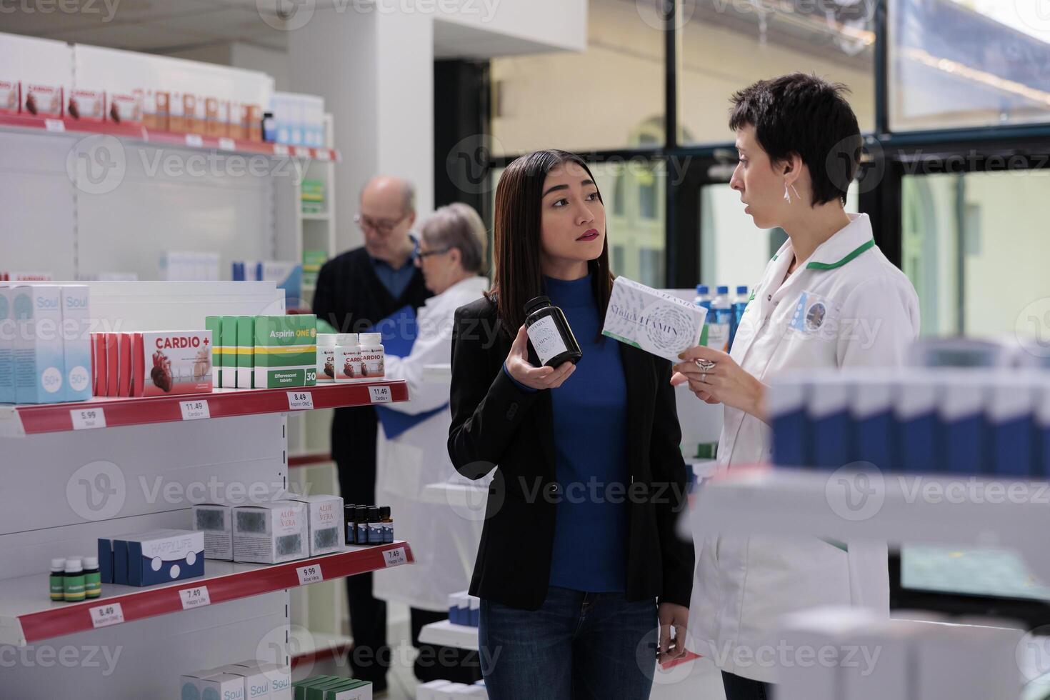 Pharmaceutical assistant and young asian woman discussing vitamin complex components in drugstore. Pharmacy employee giving assistance to customer in choosing multivitamin capsules photo