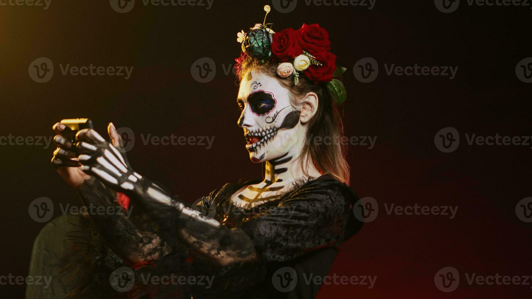 Creepy young woman playing video games on mobile phone, using mobile phone online on dios de los muertos holy tradition. Santa muerte model having fun on scary mexican festival. Handheld shot. photo