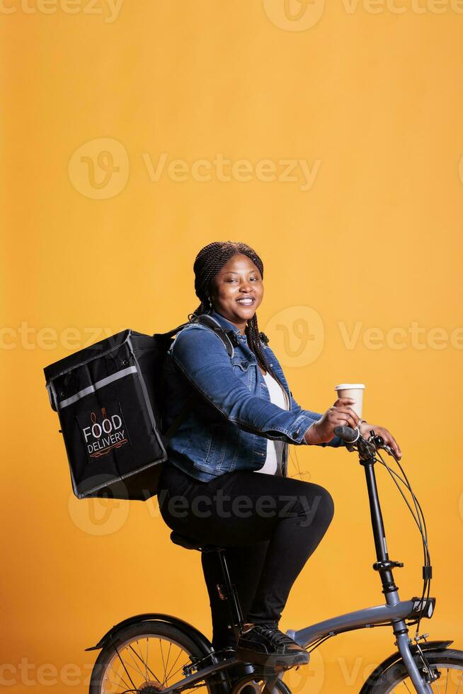 retrato de restaurante mensajero mensajero participación taza de café mientras entregando eliminar comida orden a clientes utilizando bicicleta como transporte. pizzería trabajador en pie en estudio con amarillo antecedentes. foto