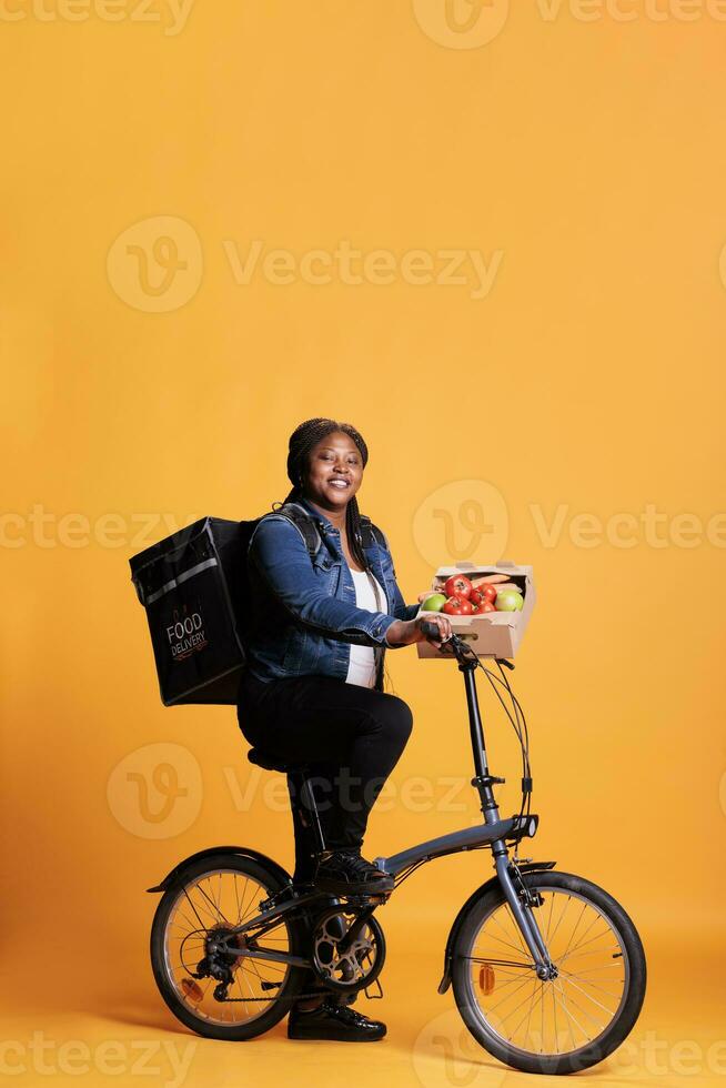 Full body of supermarket coutier holding box with healthy vegetables ready to deliver organic lunch to clients using bike as transportation. Deliverywoman carrying takeaway thermal backpack photo