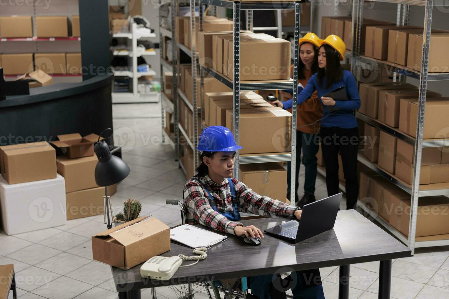 Warehouse young asian manager using wheelchair while planning logistics on laptop. Retail storehouse employee with disability analyzing customer orders checklist on computer photo