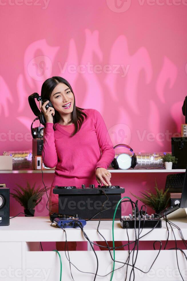 Cheerful performer with headphones playing electronic song at mixer console, standing at dj table having fun with fans at night in club. Artist performing techno music in studio over pink background photo