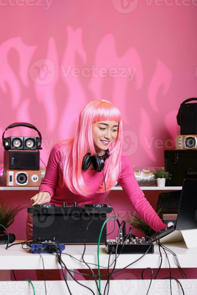 Cheeful musician standing at dj table playing electronic music with professional mixer console, enjoying to perform at night in club. Asian performer having fun with fans during techno concert photo