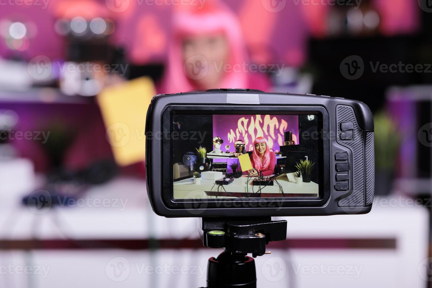 Asian vlogger holding notebook presenting product in front of camera while recording school supplies review using broadcast equipment. Content creator with pink hair live streaming photo