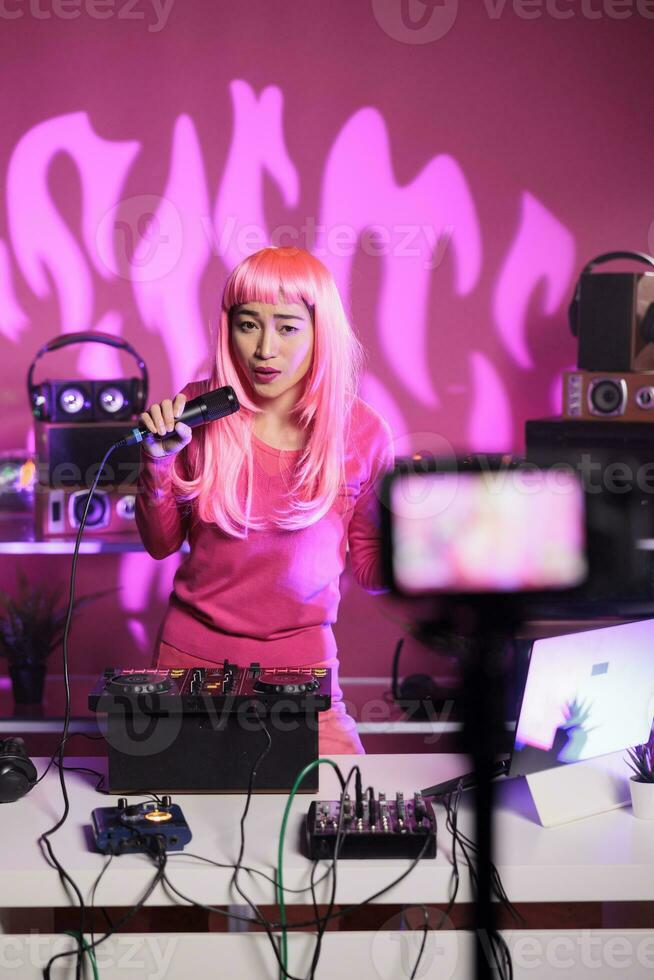 Woman standing at dj table playing techno music at mixer console while talking into microphone with fans, having fun during night time in club. Artist recording performance with professional camera photo