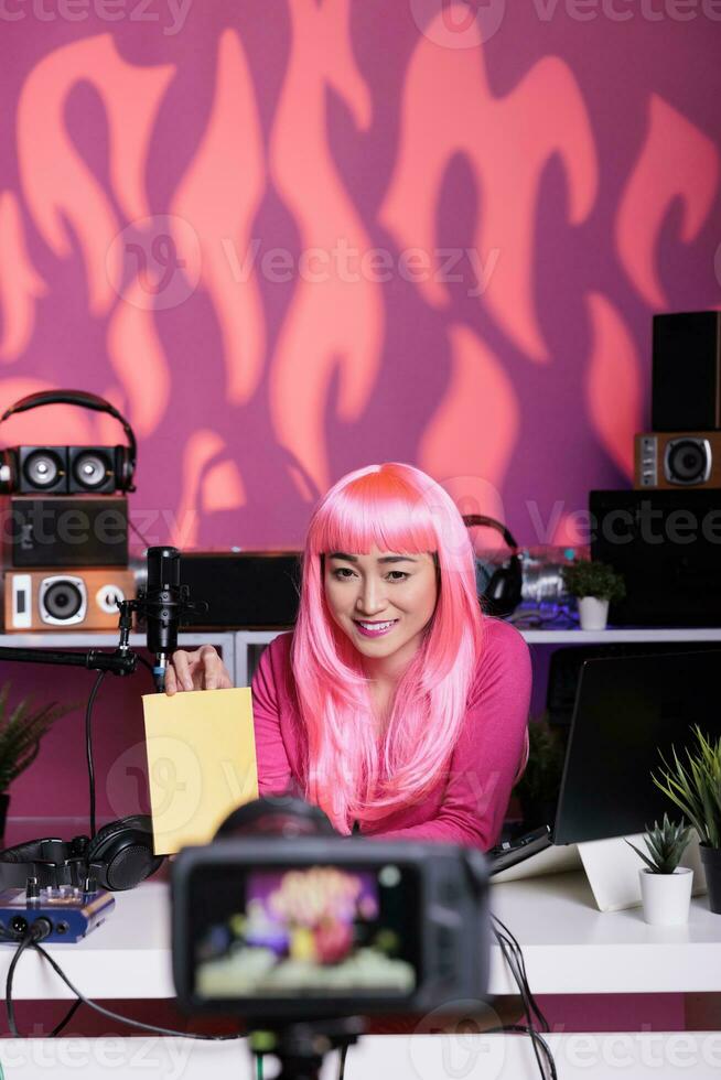 Vlogger with pink hair and tshirt showing notebook recommendation reviewing school product in front of camera while filming vlog. Asian influencer using professional broadcast equipment photo