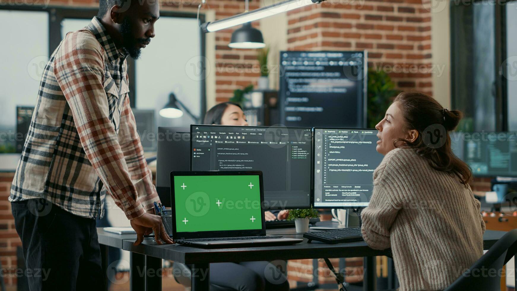 Programer working at desk with multiple computer screens and laptop with green screen chroma key mockup running code. Software developer compiling source algorithm interrupted by colleague. photo