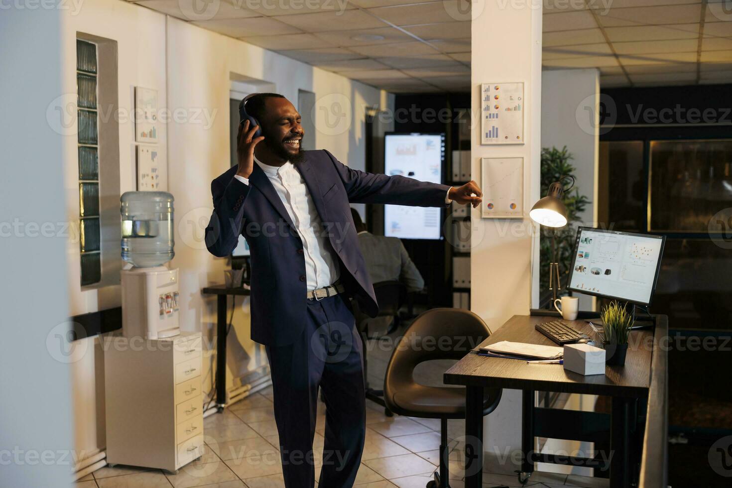 Smiling entrepreneur listening music during work break, dancing in startup office after finishing company strategy. African american businessman enjoying having fun late at night in startup office photo