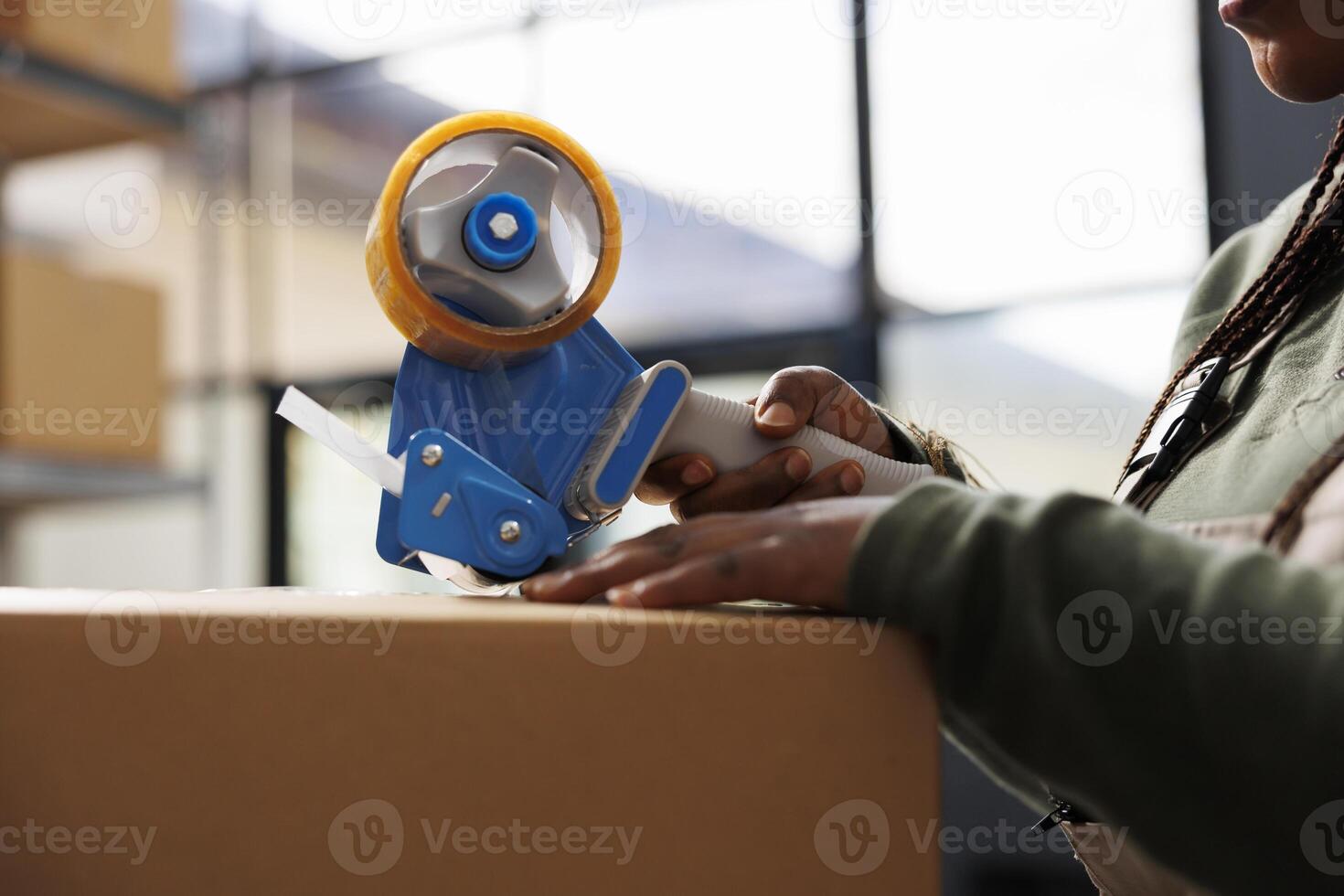 Stockroom employee preparing clients orders, putting adhesive tape on cardboard box before shipping packages. African american manager working at merchandise inventory in storage rooom. Close up photo