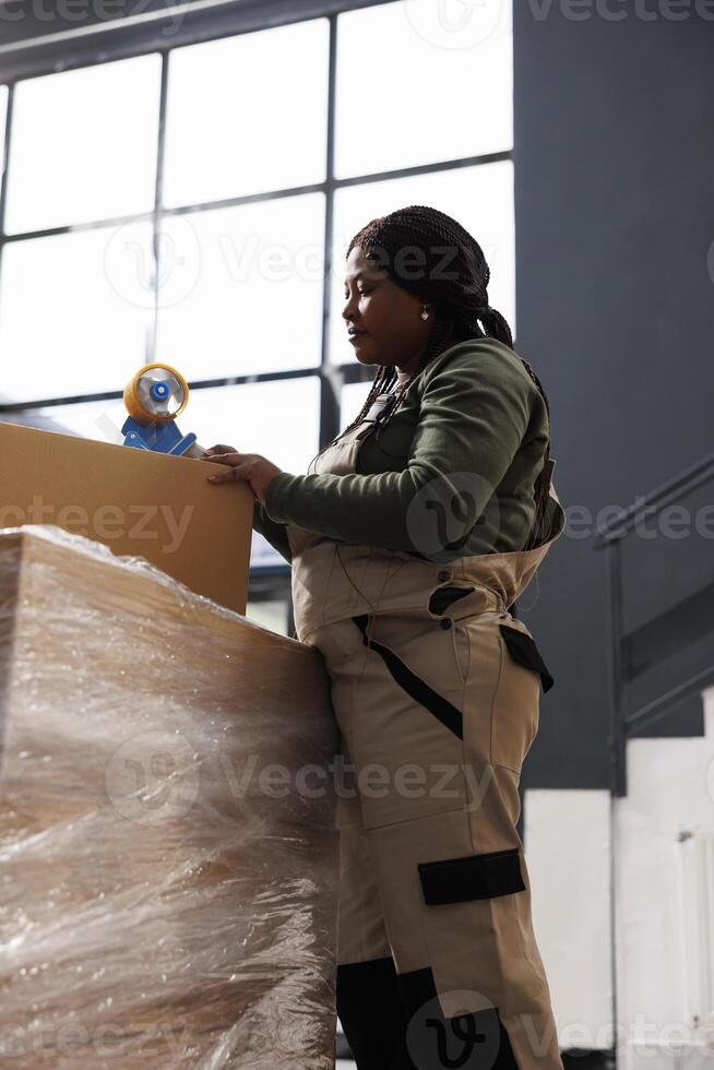 Small business employee putting adhesive tape on carton box, preparing clients packages for shipping in storehouse. Storage room supervisor wearing industrial overall working at online orders photo
