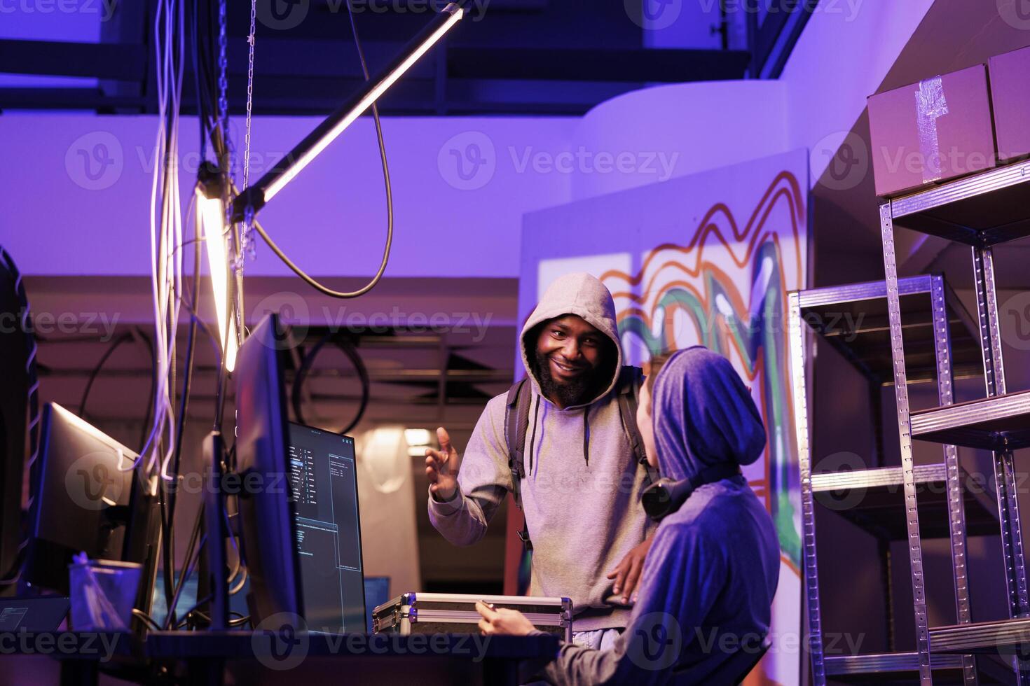 Men in hood discussing hacking plan with cheerful smile in abandoned warehouse. Happy young african american hacker explaining illegal ransomware code on computer to developer photo