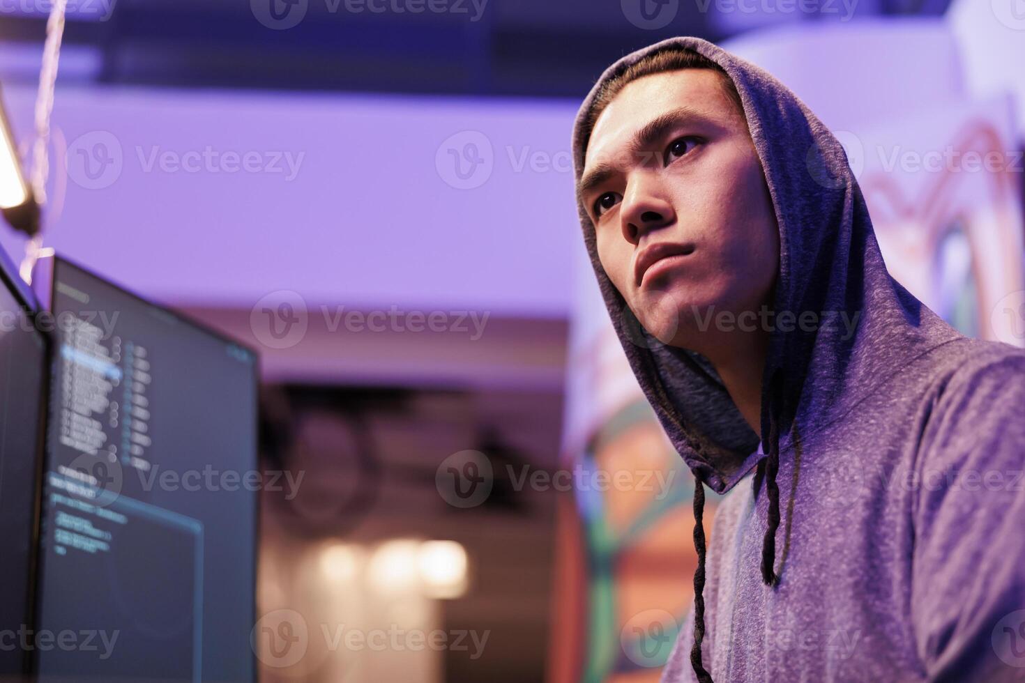 Concentrated asian criminal coding malware on computer while breaking law. Young focused hacker in hood hacking database server and using malicious software to steal data photo