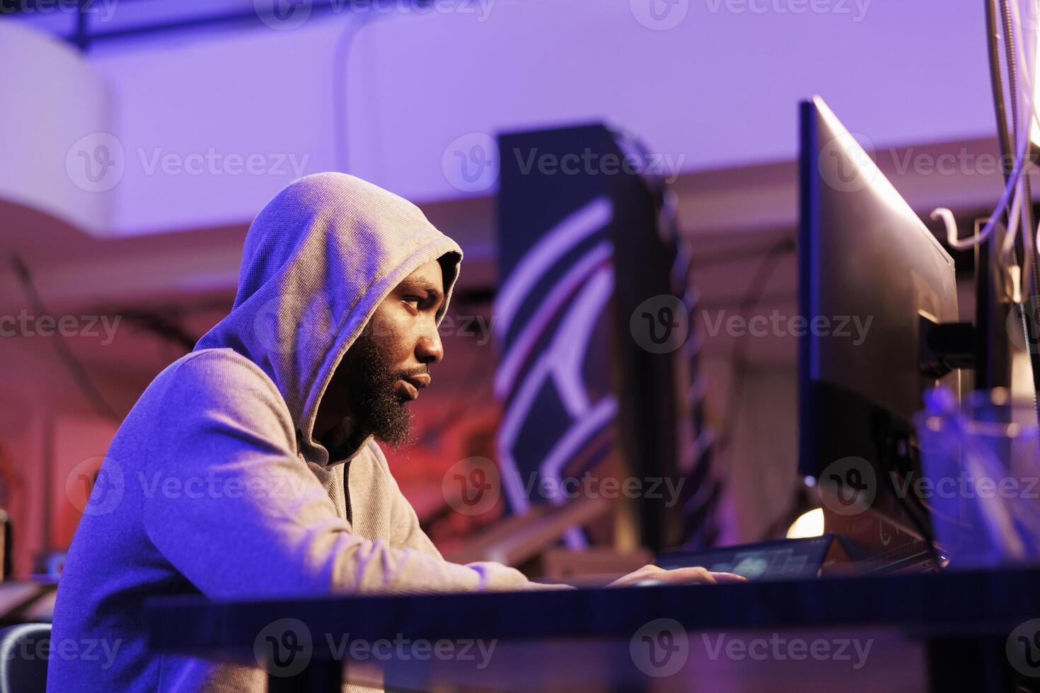 Scammer using malicious software to break into database and steal valuable information. African american hacker in hood planning phishing fraud on computer in hideout at night photo