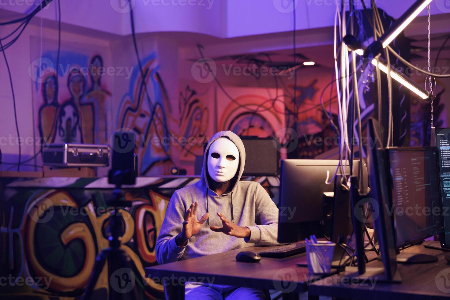 Hacker in anonymous mask live streaming illegal online activities on smartphone for dark web. Criminal recording threat on mobile phone and breaching privacy data in hideout place photo
