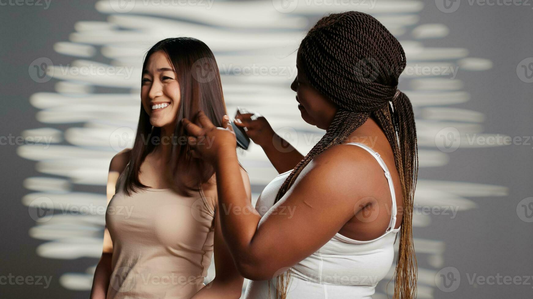 African american girl brushing hair of asian friend on camera, posing to promote friendship and wellness. Interracial women using cosmetics, advertising skincare products for ad campaign. photo