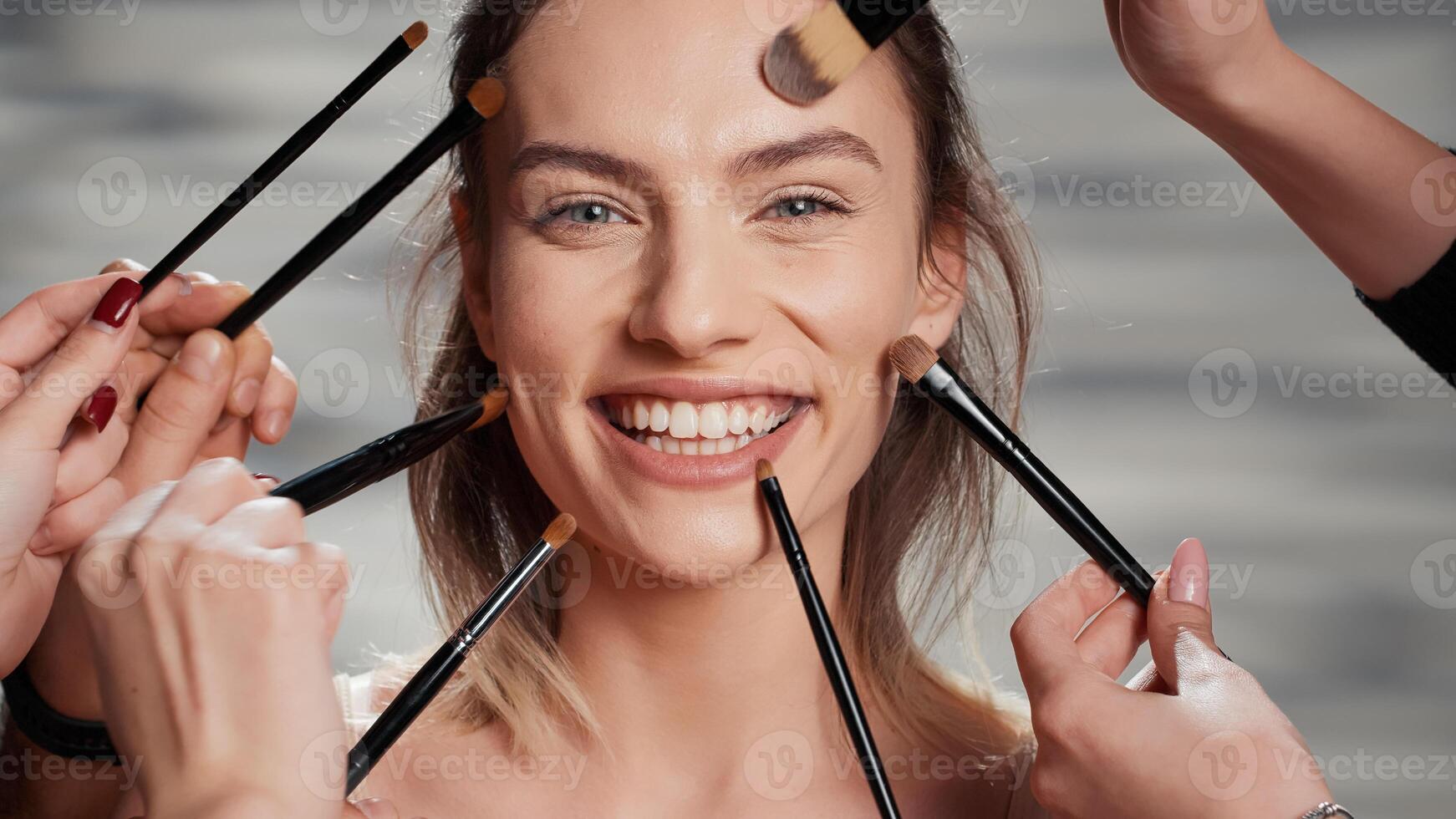 Flawless woman receiving makeover from cosmetologists, team of make up artists and specialist doing natural make up look with brushes. People preparing beauty model for ad campaign. Close up. photo