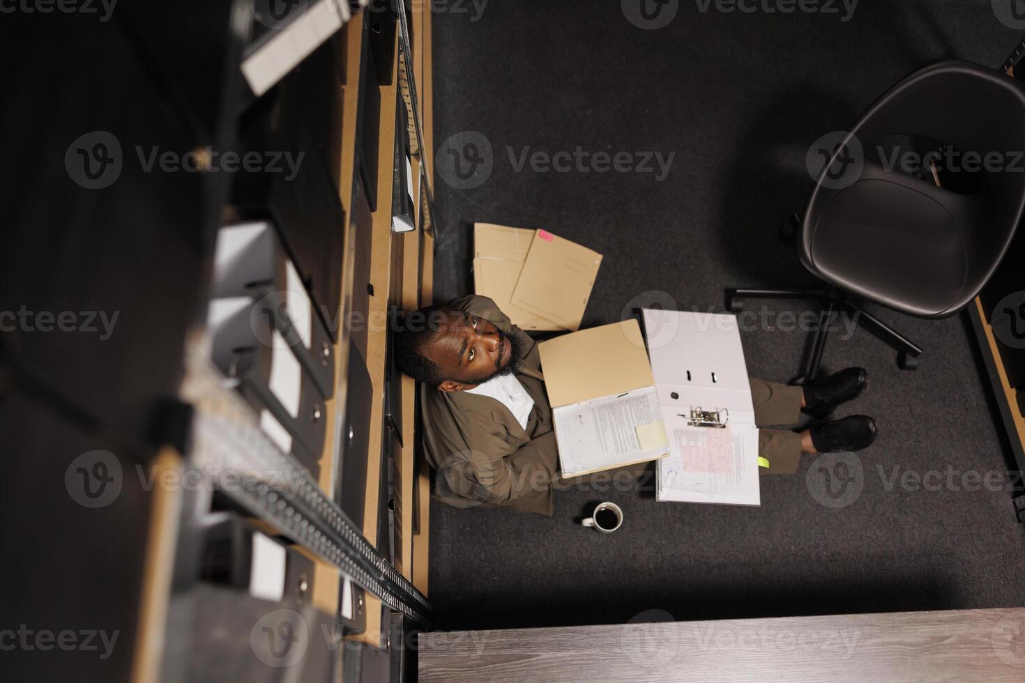 Exhausted cop sitting on floor surrounded by crime case file papers. Tired overworked investigator studying police documentation in office late and looking upwards top view photo