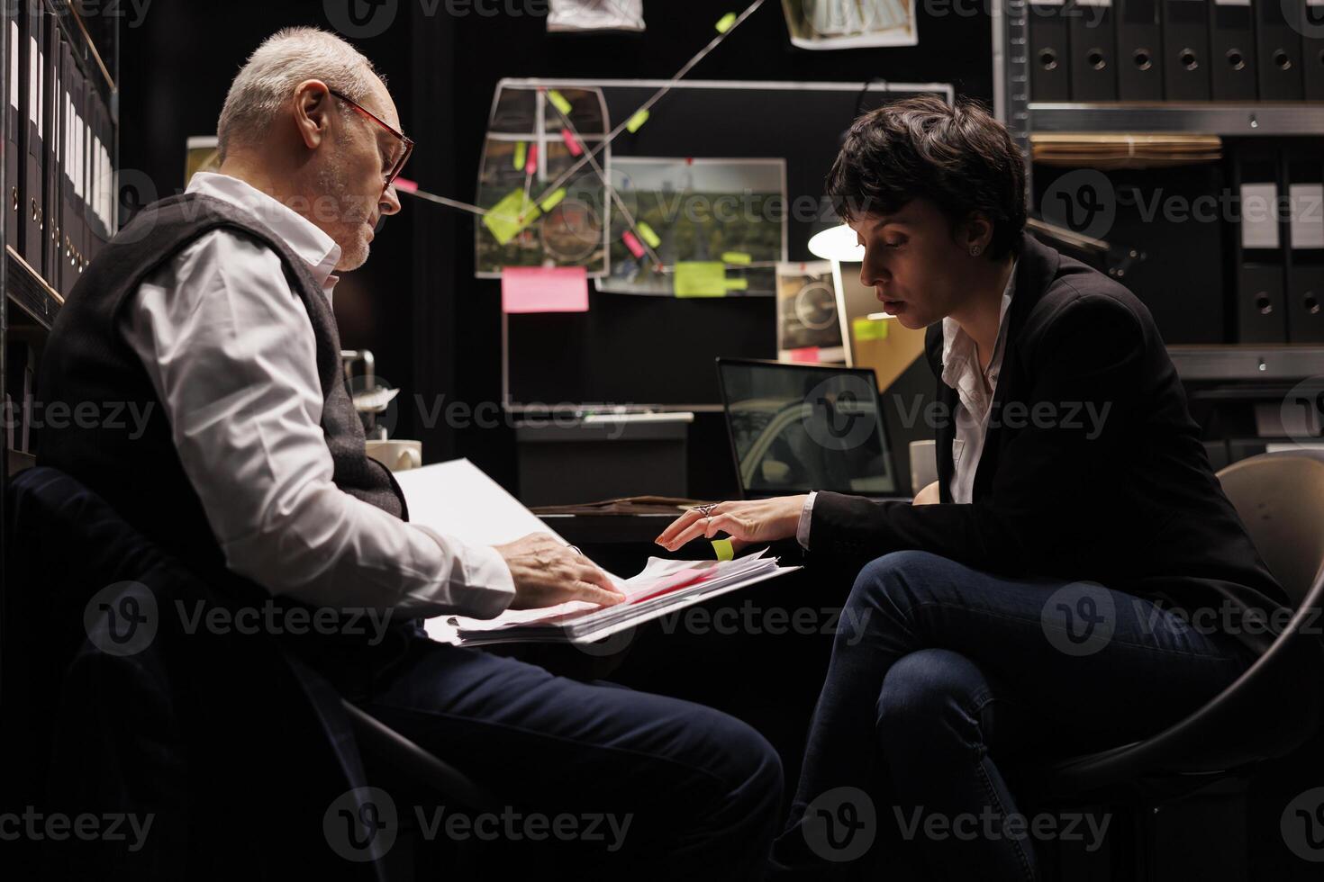 Old investigator showing confidential files to woman agent, discussing crime scene evidence late at night in dark room. Caucasian private detectives working at solution to solve criminal case photo