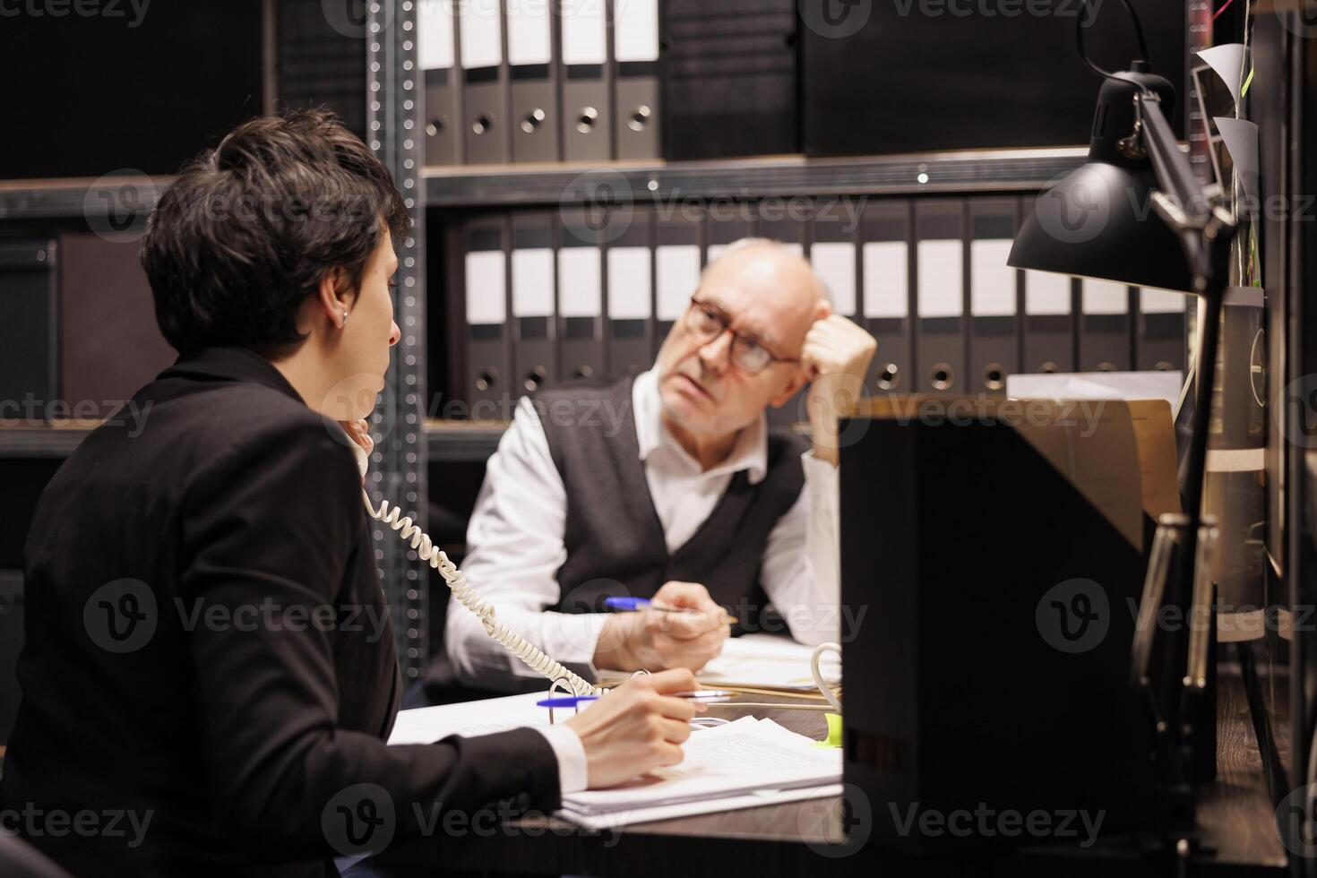 Woman private detective talking at landline phone, discussing criminology report with remote police officer. Senior investigator analyzing crime scene evidence, working overtime in arhive room photo