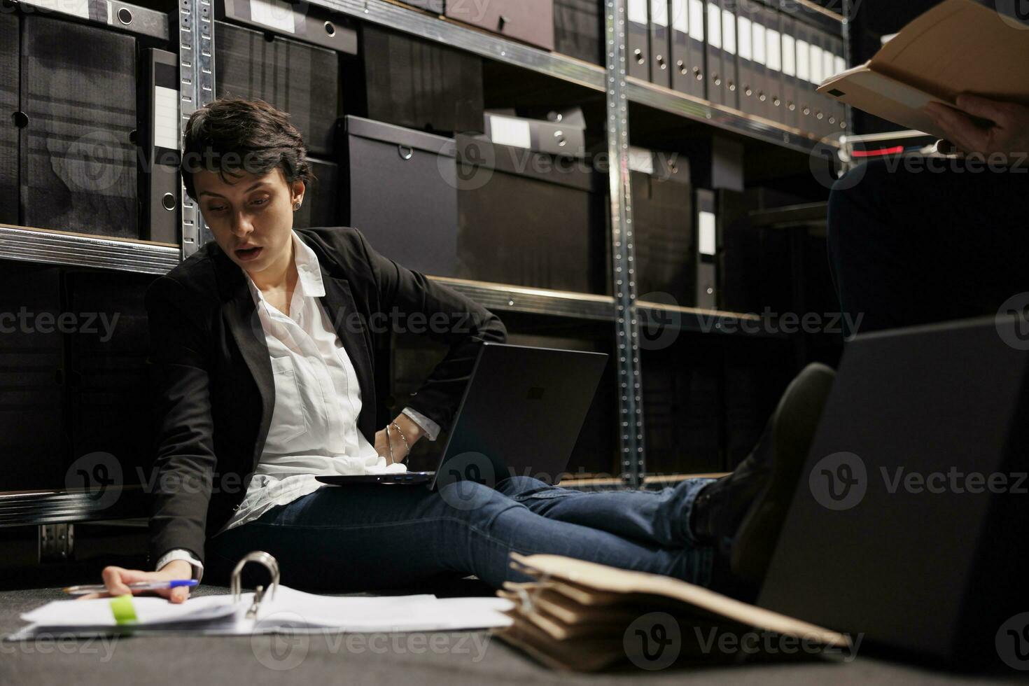 Overworked inspector sitting on floor in arhive room, analyzing criminology report. Private detectives working overhours at criminal case, checking crime scene evidence trying to catch suspect photo