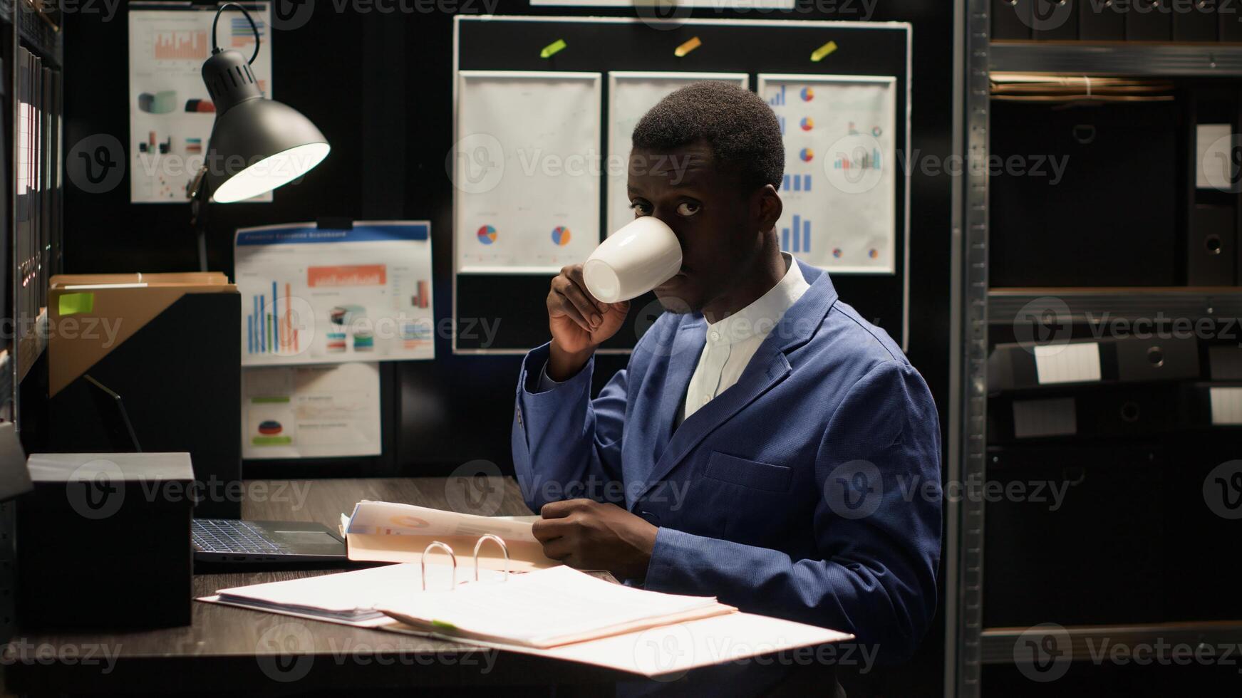 Male inspector with expertise in criminal investigation surrounded by files and evidence, looking at camera with confident smile. Police officer analyzing clues in incident room while drinking coffee. photo