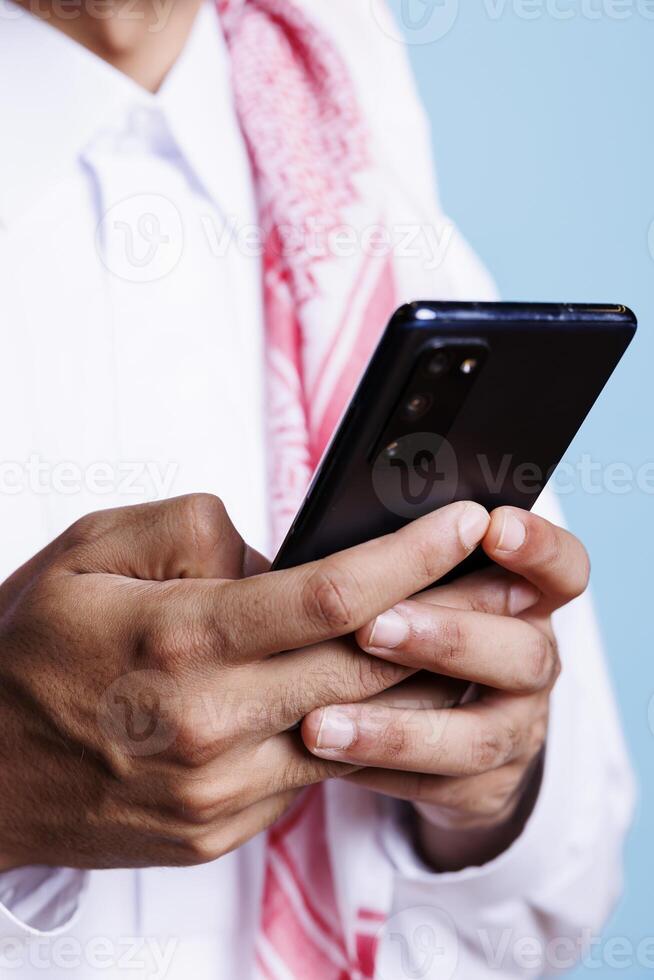 Muslim man holding mobile phone in arms, sending online message while using chatting app closeup. Arab in traditional thobe using smartphone and scrolling social media page photo