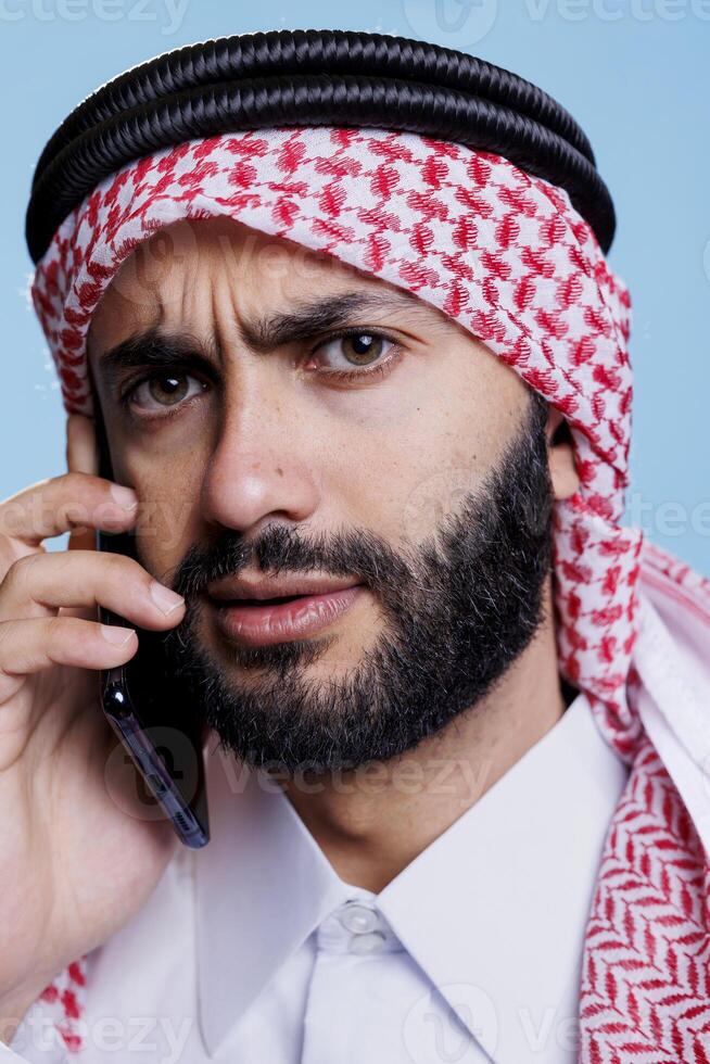 Arab man frowning eyebrows and looking at camera with confused expression while chatting on smartphone. Muslim person wearing ghutra speaking on mobile phone closeup portrait photo