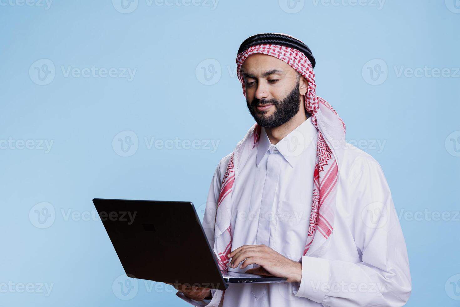 Muslim man wearing traditional thobe and ghutra headscarf holding laptop and broswing social media. Arab person dressed in islamic clothes using portable computer and checking internet page photo