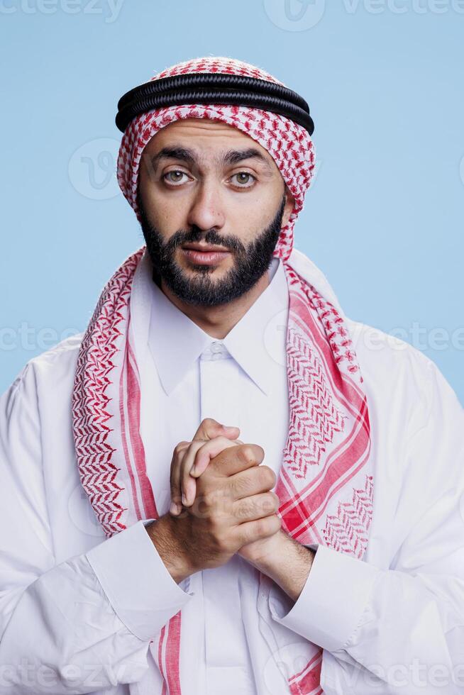 Muslim man praying while showing faith and belief and looking at camera with hopeful expression. Arab prayer posing with folded hands while praising god studio portrait on blue background photo