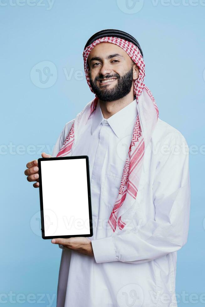 Smiling muslim man showing digital tablet with empty white screen and looking at camera with cheerful expression. Happy arab advertiser holding blank touchscreen portrait photo