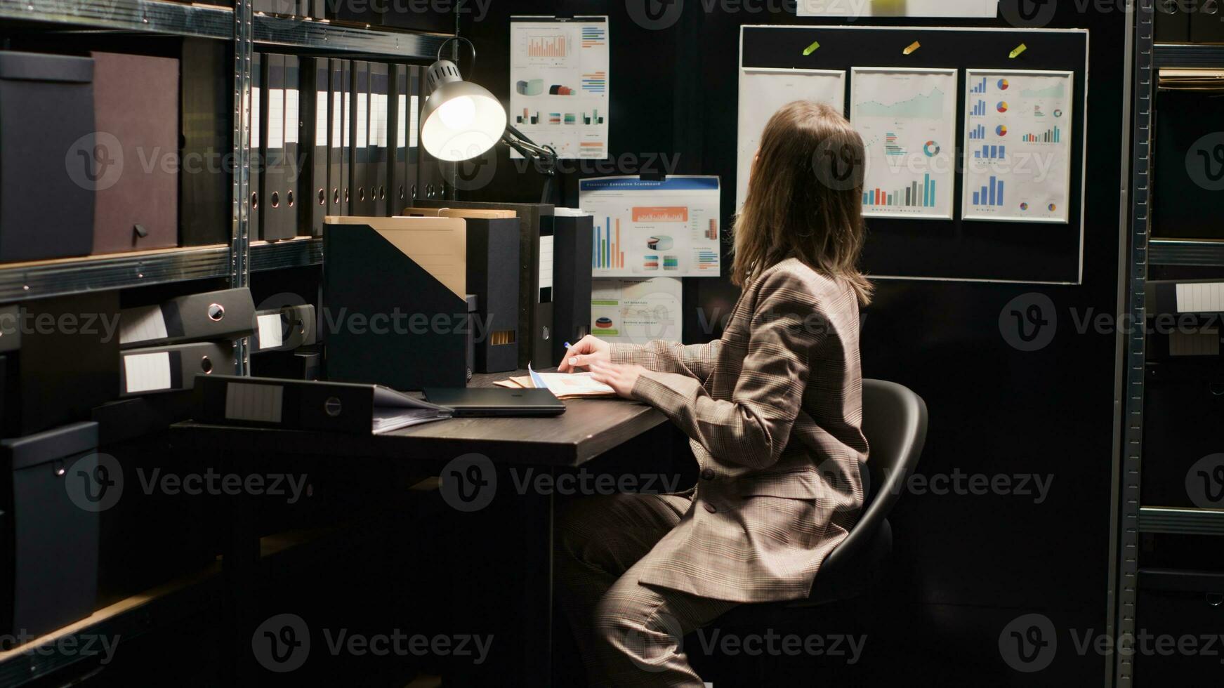 Caucasian private investigator sits at desk with minicomputer and reviews suspect case material. Female law officer with expertise in surveillance, enters discreet office space holding laptop bag. photo