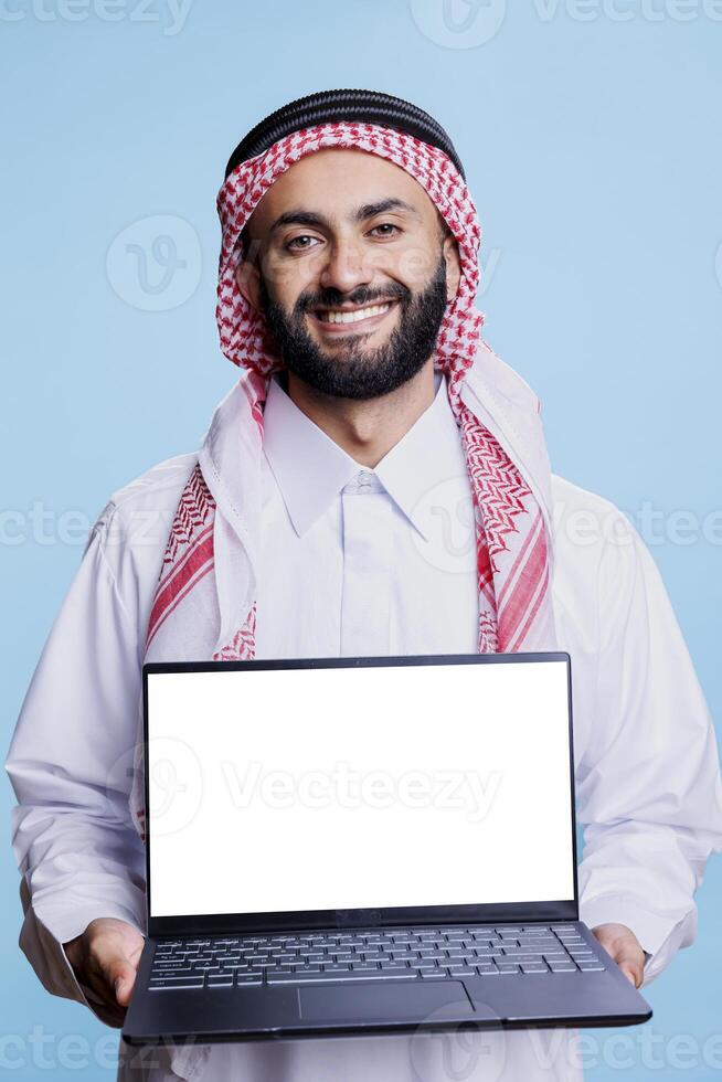 Smiling muslim man showing laptop with blank screen mockup for software advertising portrait. Arab presenting computer with empty display and looking at camera with cheerful expression photo