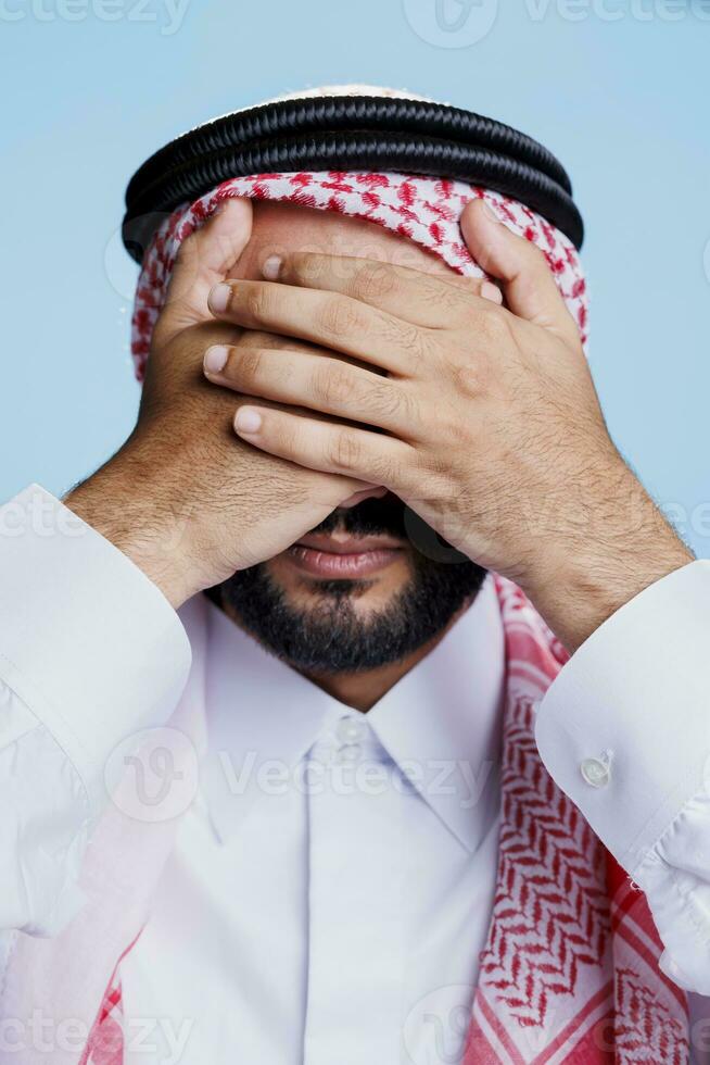 Man dressed in islamic clothes closing eyes with hands while showing see no evil three wise monkeys sign. Young muslim person showcasing eyesight problem concept closeup in studio photo