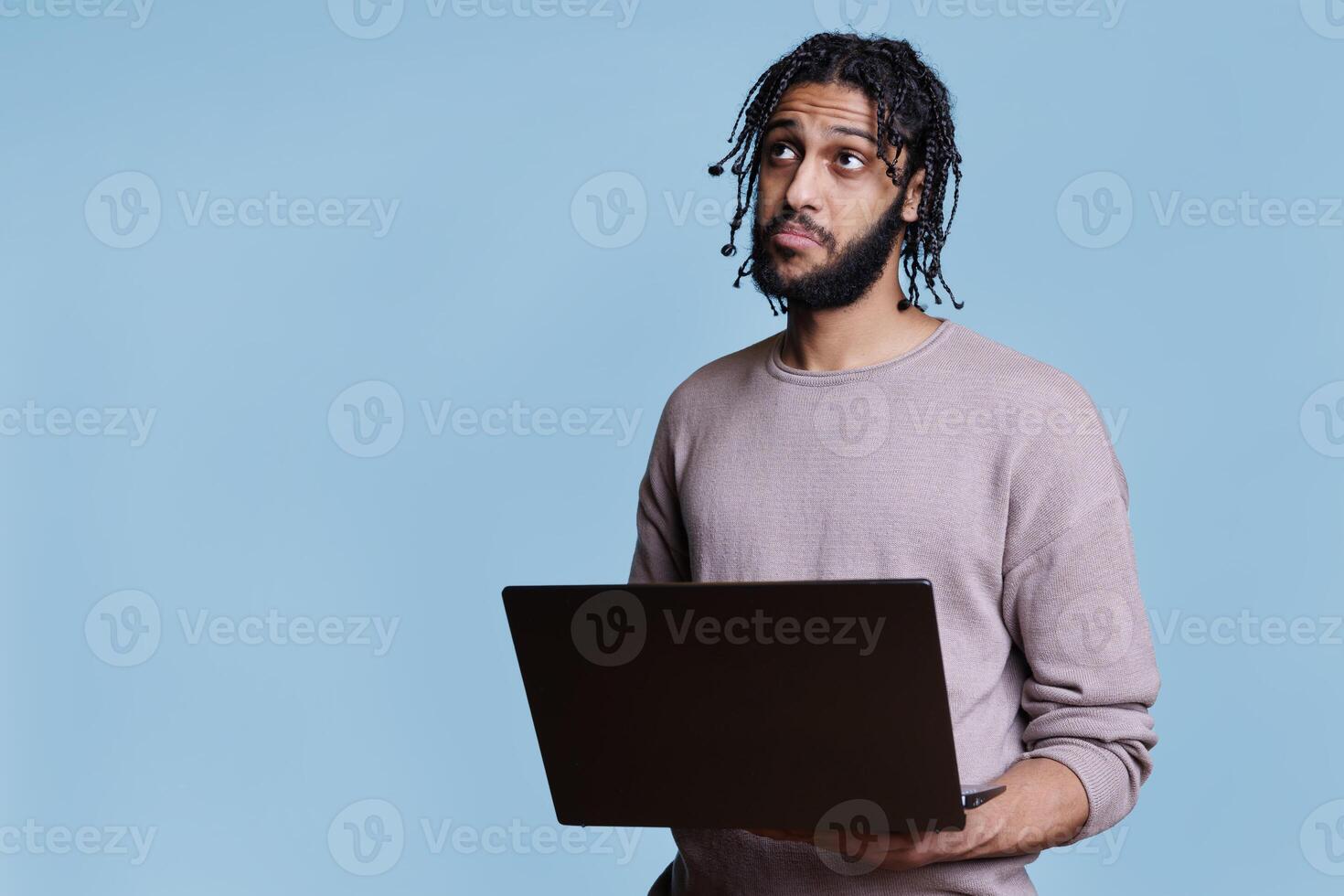 Pensive arab man thinking about business report while holding laptop and looking up. Thoughtful person with puzzled facial expression analyzing start up project on portable computer photo