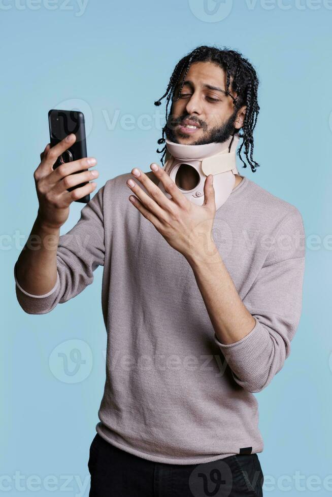 Arab patient with cervical collar having online consultation with doctor on smartphone. Man wearing neck brace talking with general practitioner on smartphone about trauma photo