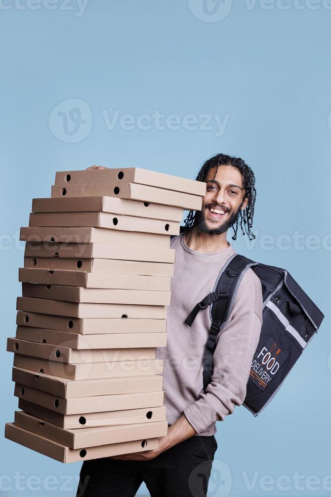 Carefree arab deliveryman carrying big pizza boxes pile and looking at camera with cheerful expression. Smiling restaurant delivery service courier standing huge stack of fast food packages photo