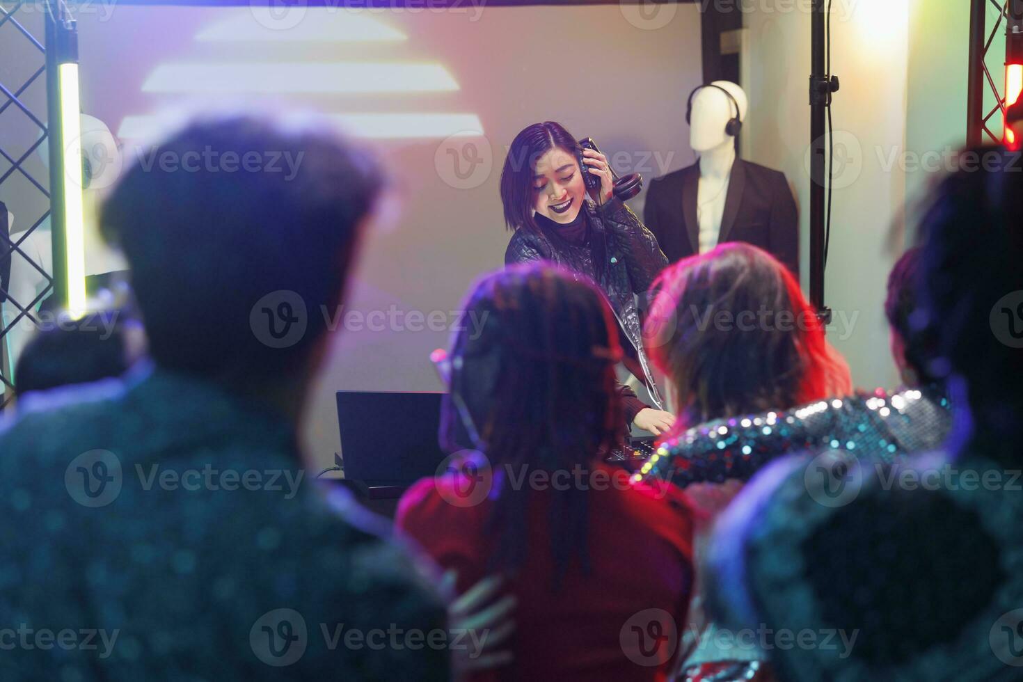 Crowd watching musician performance while attending discotheque party in nightclub. Young asian woman dj playing using headphones and digital controller on stage with spotlights in club photo