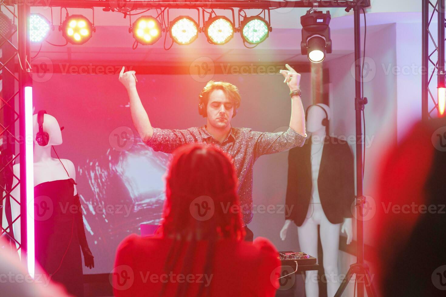 Young man dj in headphones standing on stage with spotlights at nightclub concert. Musician with closed eyes raising hands while enjoying electronic music at disco party in club photo