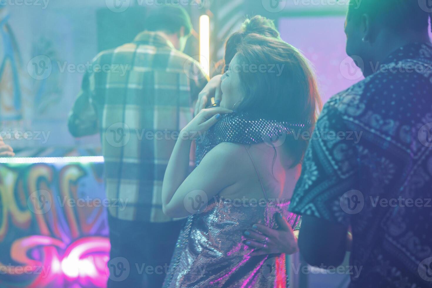 Women hugging in nightclub while attending discotheque party. Girlfriends embracing on dancefloor with spotlights while dancing and enjoying nightlife entertainment in club at night photo