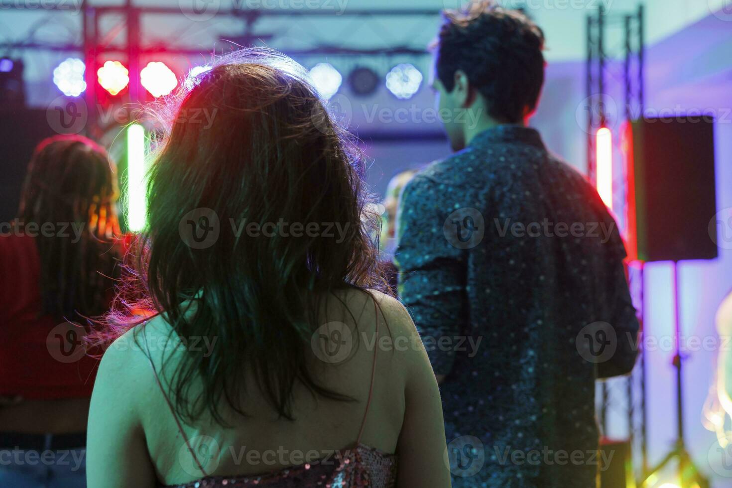 People standing on dancefloor while attending concert in nightclub with spotlights. Crowd partying, celebrating and dancing at discotheque while enjoying music in club back view photo