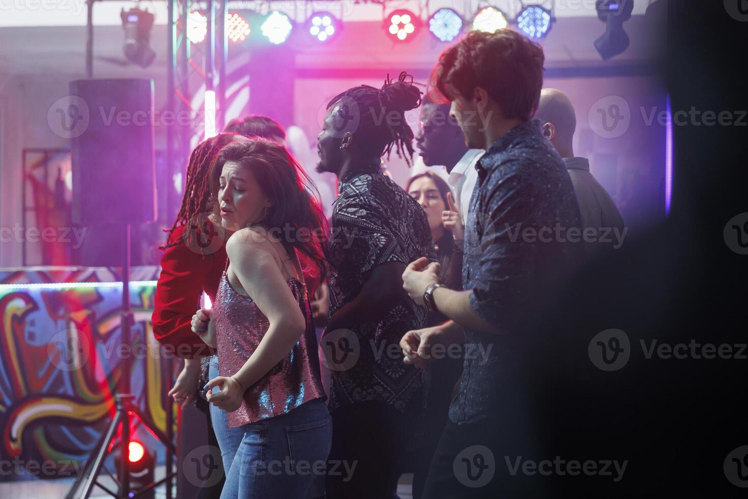 Young woman making passionate dance moves while clubbing with friends at discotheque. Crowd partying on vibrant dancefloor while attending disco party with live music in nightclub photo