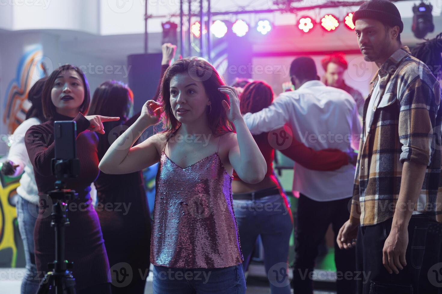 Young woman taking selfie on smartphone with tripod while clubbing. Caucasian girl partying at nightclub discotheque and live streaming festival event in social media using mobile phone photo