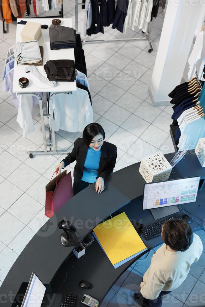 Smiling asian woman talking with cashier while making purchase at clothes store checkout desk top view. Shopping center boutique satisfied customer standing at cash register photo