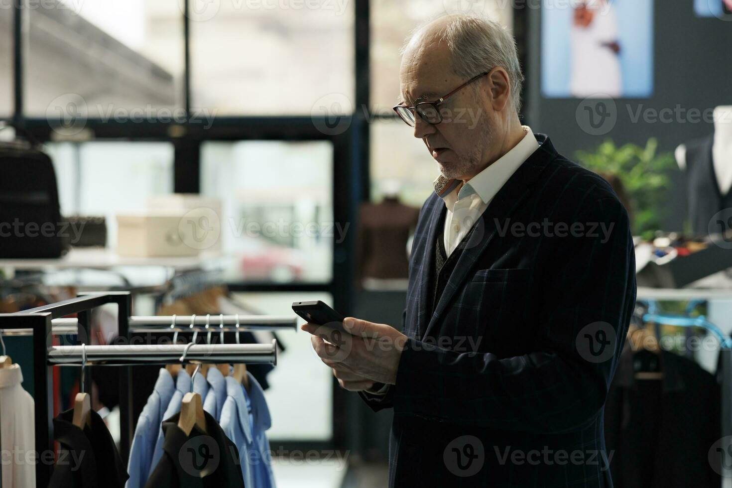 Elderly man shopping for casual wear in showroom, messaging wife using mobile phone. Senior man wearing formal suit buying fashionable clothes and trendy accessories in modern boutique photo