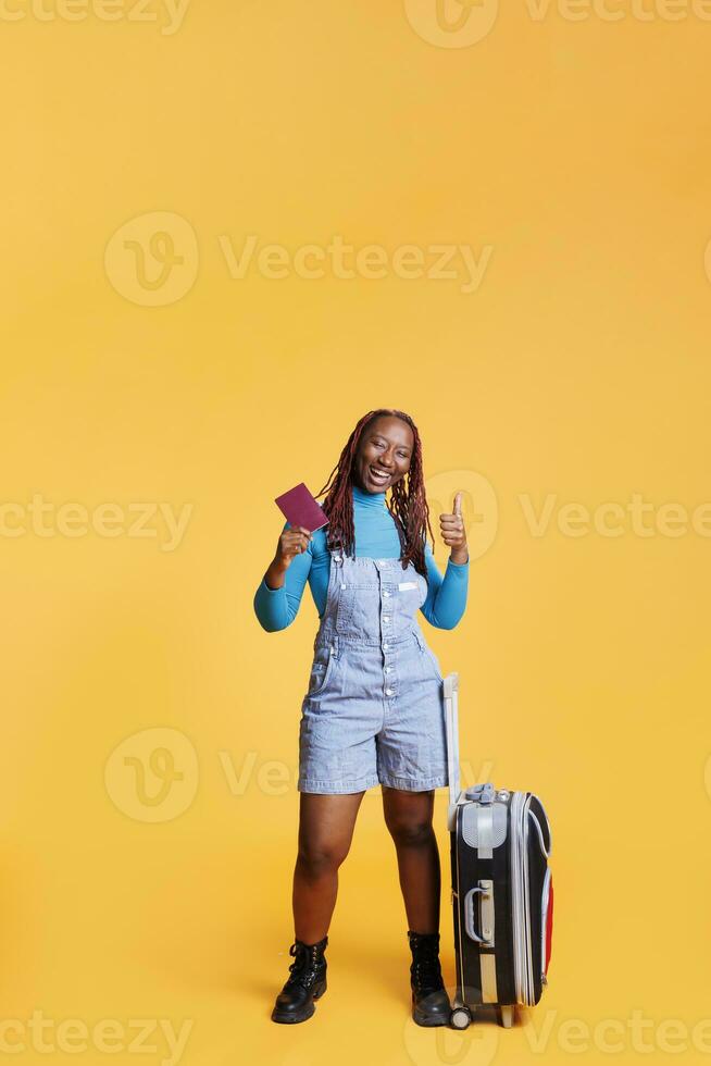 viajero dando pulgares arriba en estudio, que lleva carretilla pantalones y pasaporte antes de volador en el extranjero. alegre mujer disfrutando urbano aventuras con identificación aprobar, internacional destino. foto