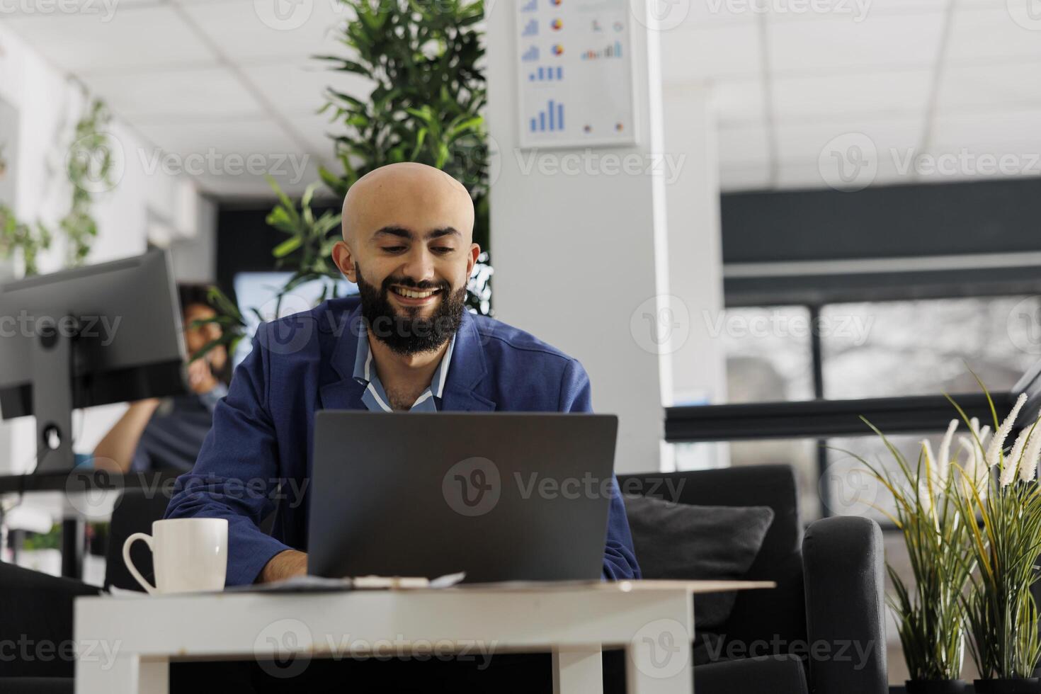 Smiling arab businessman working on laptop in business office while sitting in couch. Start up company executive manager analyzing successful project sales on portable computer in coworking space photo