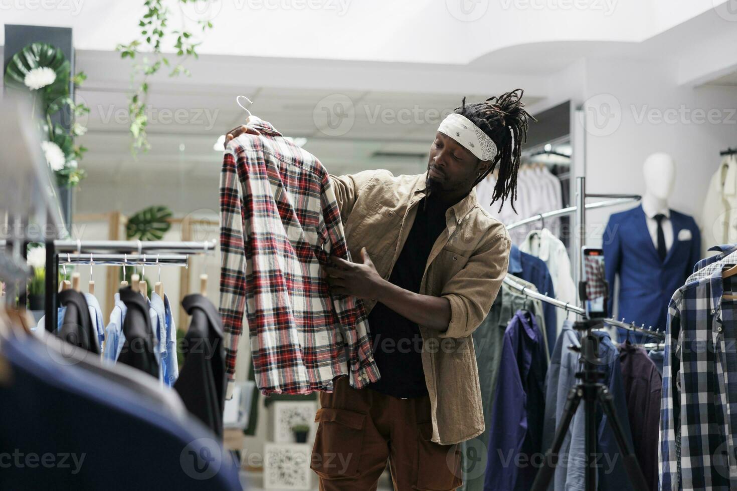 African american man promoting clothing store new collection on social media, filming video with mobile phone. Fashion blogger holding shirt and speaking about trendy style on smartphone camera photo