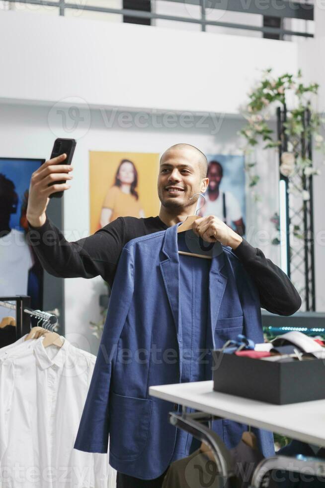 Arab blogger taking selfie on smartphone, creating content for fashion blog in clothing store. Influencer recording video on mobile phone while promoting clothing store offerings photo