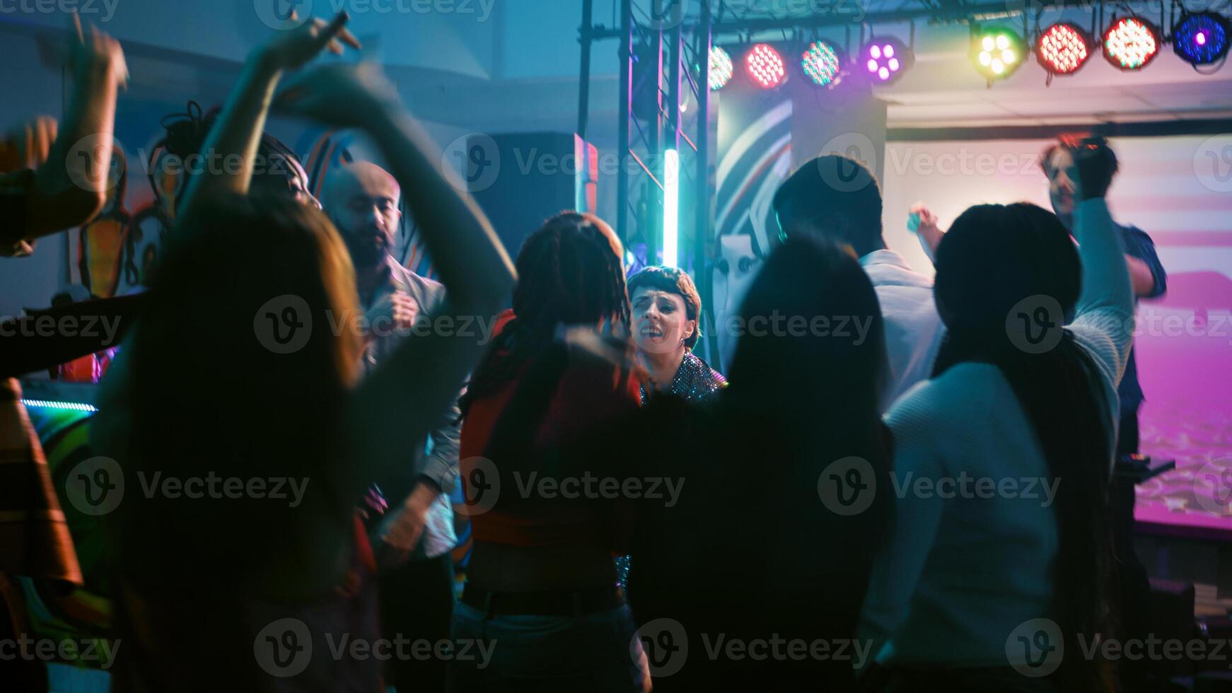 joven adultos bailando en electrónico música, teniendo divertido juntos con DJ en escenario. alegre hombres y mujer saltando en danza piso debajo disco luces, disfrutando danza fiesta. Mano disparo. foto