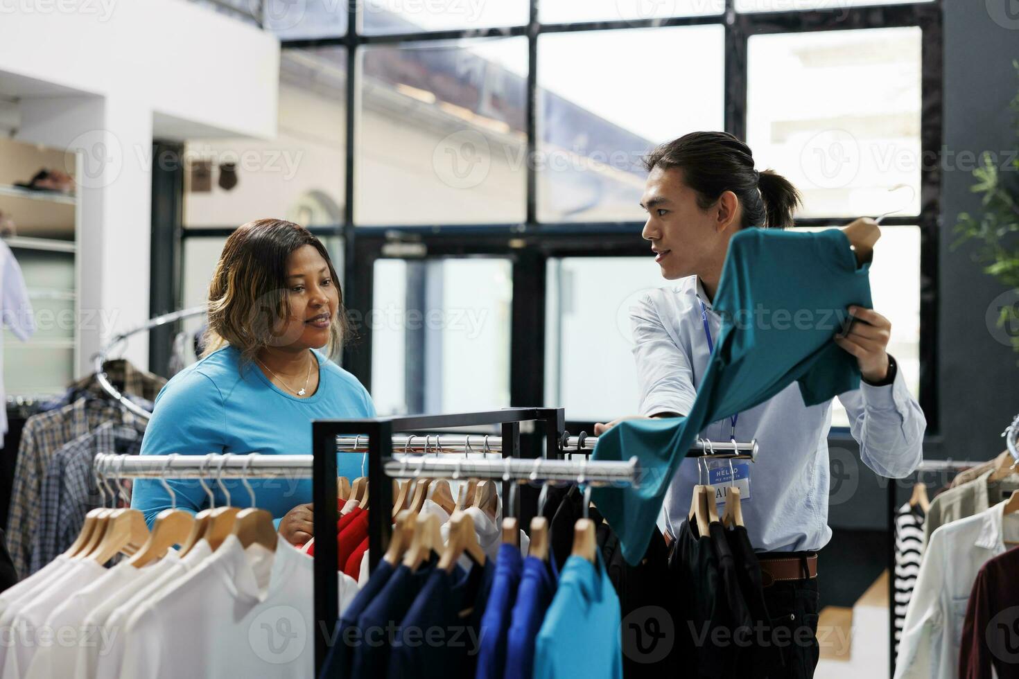 African american customer discussing clothes fabric, looking at racks full with fashionable merchandise in clothing store. Stylish woman choosing to buy casual wear in modern boutique photo